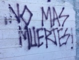 Graffiti text on a textured white brick wall reads 'NO MAS MUERTES!' in bold, dark spray paint. The background shows visible brick lines and some weathering.