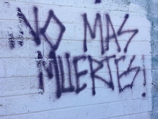 Graffiti text on a textured white brick wall reads 'NO MAS MUERTES!' in bold, dark spray paint. The background shows visible brick lines and some weathering.