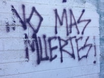 Graffiti text on a textured white brick wall reads 'NO MAS MUERTES!' in bold, dark spray paint. The background shows visible brick lines and some weathering.