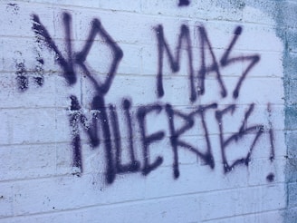 Graffiti text on a textured white brick wall reads 'NO MAS MUERTES!' in bold, dark spray paint. The background shows visible brick lines and some weathering.