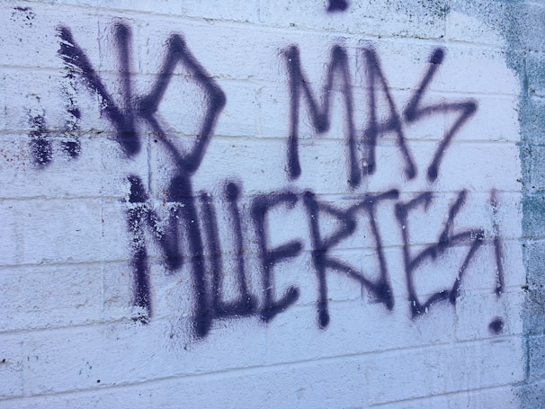 Graffiti text on a textured white brick wall reads 'NO MAS MUERTES!' in bold, dark spray paint. The background shows visible brick lines and some weathering.