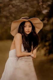 A young woman with long dark hair wearing a wide-brimmed hat leans against a stone railing. Her white dress contrasts softly with the blurred, green and brown background, creating an elegant and serene atmosphere.