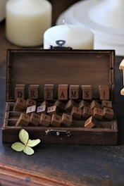 A rustic wooden box filled with letter tiles spelling out the word 'DREAMS' along with other scattered letters. Two unlit, white candles and a dried, pale green leaf accompany the scene, adding a vintage and contemplative atmosphere.