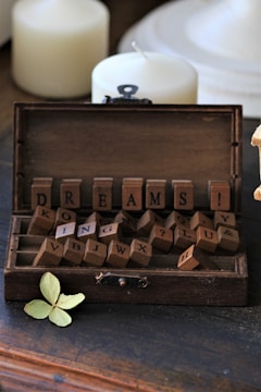 A rustic wooden box filled with letter tiles spelling out the word 'DREAMS' along with other scattered letters. Two unlit, white candles and a dried, pale green leaf accompany the scene, adding a vintage and contemplative atmosphere.