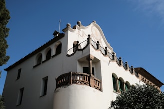 A historic building with a decorative facade, featuring arched windows and intricate wrought-iron details. The architecture showcases traditional design elements with a predominantly white exterior, set against a clear blue sky.