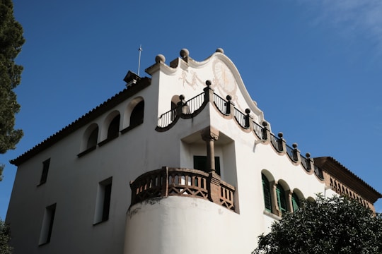 A historic building with a decorative facade, featuring arched windows and intricate wrought-iron details. The architecture showcases traditional design elements with a predominantly white exterior, set against a clear blue sky.