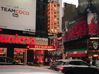 A bustling urban street scene with bright lights and large signs. Prominent advertisements include a sign for 'TEAMCOCO' and a large neon sign for 'Juniors' restaurant, established in 1950. The area is filled with vibrant colors and multiple signs advertising food and entertainment venues. Cars are visible in the foreground, and the street is alive with various forms of signage and visual noise.