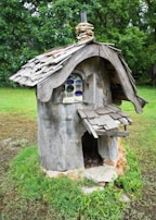A whimsical, rustic structure resembling a small house made from tree bark and wood. The roof is constructed with uneven wooden shingles, and there is a tiny chimney made from stacked stone. A window with colorful, round panes adds a charming detail. The structure stands on a grassy area surrounded by lush green foliage.