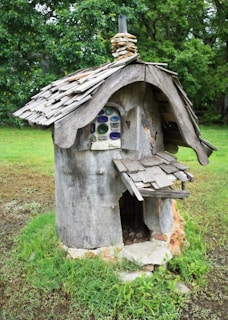 A whimsical, rustic structure resembling a small house made from tree bark and wood. The roof is constructed with uneven wooden shingles, and there is a tiny chimney made from stacked stone. A window with colorful, round panes adds a charming detail. The structure stands on a grassy area surrounded by lush green foliage.