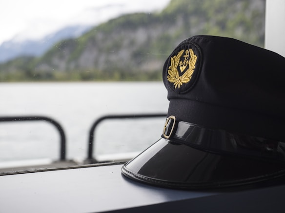 A black captain's hat with a gold emblem featuring an anchor and laurel leaves sits on a surface. In the background, there is a body of water and blurred green hills, suggesting a maritime setting.