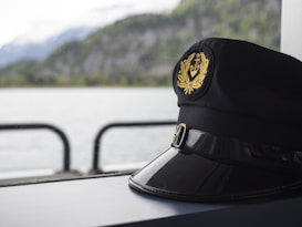 A black captain's hat with a gold emblem featuring an anchor and laurel leaves sits on a surface. In the background, there is a body of water and blurred green hills, suggesting a maritime setting.