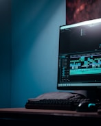 A computer monitor displays video editing software with a timeline and various clips. The setup includes a keyboard and a mouse, situated on a desk in a room with a bluish hue.