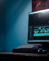 A computer monitor displays video editing software with a timeline and various clips. The setup includes a keyboard and a mouse, situated on a desk in a room with a bluish hue.
