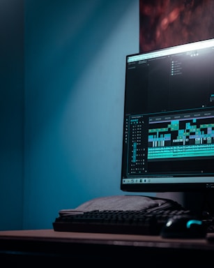 A computer monitor displays video editing software with a timeline and various clips. The setup includes a keyboard and a mouse, situated on a desk in a room with a bluish hue.