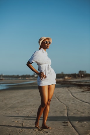 A woman stands confidently on a sandy beach wearing a white dress, sunglasses, and a wide-brimmed hat. The sky is clear and blue, and the beach stretches into the distance with faintly visible trees and water along the shore.