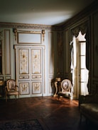 A luxurious, ornately decorated room with intricate wall panels and gold detailing. The space features elegant chairs with floral upholstery, a richly patterned wooden floor, and a window with heavy drapes letting in soft light.