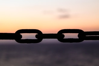 A silhouetted chain is prominently featured against a gradient sky as the sun sets, creating a serene backdrop. The links of the chain appear dark and distinct, set against the warm hues of the sunset.