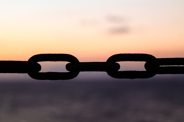 A silhouetted chain is prominently featured against a gradient sky as the sun sets, creating a serene backdrop. The links of the chain appear dark and distinct, set against the warm hues of the sunset.
