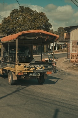A small truck with a canvas cover is driving down a rural road lined with a few buildings and trees. The truck has visible writing on the back and is casting shadows on the road. The scene includes some people on a motorcycle nearby, and the sunlight gives the image a warm, late afternoon appearance.