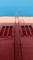 A vertical black metal ladder is attached to a red wall, leading upwards with a bright blue sky in the background. The wall has a series of rectangular indented sections, and the ladder casts a shadow on the wall.