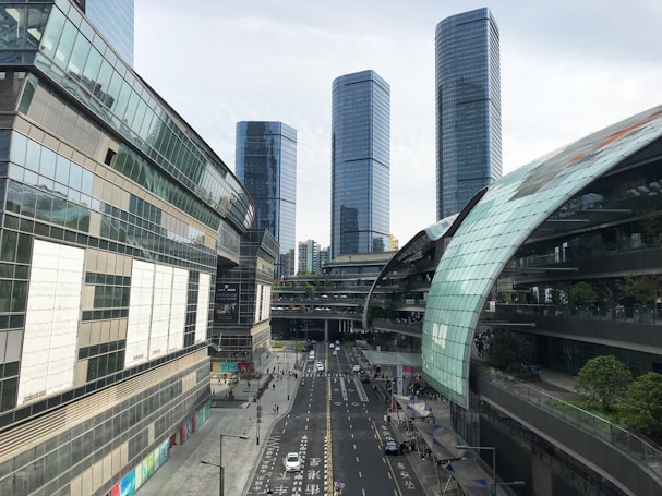 A modern cityscape with tall, sleek skyscrapers made of glass and steel. The buildings have a reflective surface, and there is a road running through the middle with cars and pedestrians. The sky is overcast, and various shades of gray and green are visible on the buildings.
