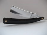 A close-up view of a straight razor, featuring a shiny metallic blade and a black handle. The blade has the text 'Wade & Butcher For Barber's Use' engraved on it. The razor is resting on a white surface with a plain, light-colored background.