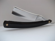 A close-up view of a straight razor, featuring a shiny metallic blade and a black handle. The blade has the text 'Wade & Butcher For Barber's Use' engraved on it. The razor is resting on a white surface with a plain, light-colored background.