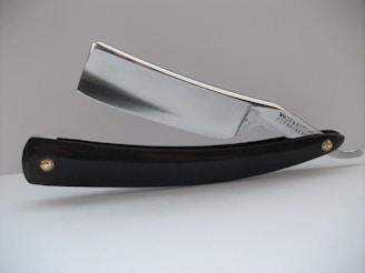 A close-up view of a straight razor, featuring a shiny metallic blade and a black handle. The blade has the text 'Wade & Butcher For Barber's Use' engraved on it. The razor is resting on a white surface with a plain, light-colored background.