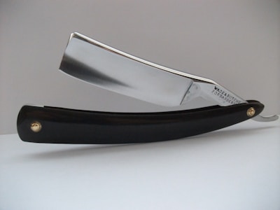 A close-up view of a straight razor, featuring a shiny metallic blade and a black handle. The blade has the text 'Wade & Butcher For Barber's Use' engraved on it. The razor is resting on a white surface with a plain, light-colored background.