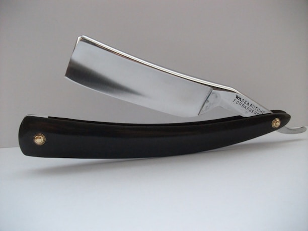 A close-up view of a straight razor, featuring a shiny metallic blade and a black handle. The blade has the text 'Wade & Butcher For Barber's Use' engraved on it. The razor is resting on a white surface with a plain, light-colored background.