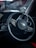 A close-up view of a car steering wheel featuring the Kia logo in the center. The dashboard and part of the interior, with some controls and dials visible, are shown in low light. The background appears blurred, hinting at a street or outdoor setting.