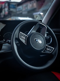 A close-up view of a car steering wheel featuring the Kia logo in the center. The dashboard and part of the interior, with some controls and dials visible, are shown in low light. The background appears blurred, hinting at a street or outdoor setting.