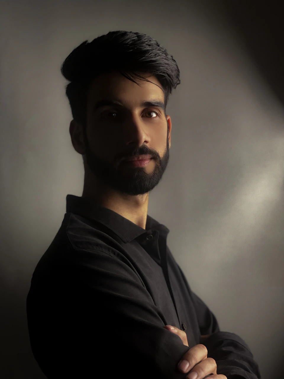 Young man with dark hair and beard looks directly at camera with crossed arms