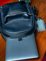 A black leather backpack is placed on a wooden surface. A silver laptop, partially visible, is tucked underneath the backpack. The zipper on the backpack is closed, and there is a tag attached to it with the brand name on display. In the background, some greenery can be seen.