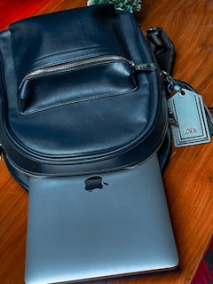 A black leather backpack is placed on a wooden surface. A silver laptop, partially visible, is tucked underneath the backpack. The zipper on the backpack is closed, and there is a tag attached to it with the brand name on display. In the background, some greenery can be seen.