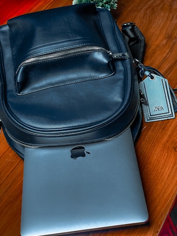 A black leather backpack is placed on a wooden surface. A silver laptop, partially visible, is tucked underneath the backpack. The zipper on the backpack is closed, and there is a tag attached to it with the brand name on display. In the background, some greenery can be seen.
