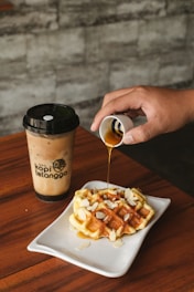 A wooden table holds a white plate with a waffle topped with almond slices and drizzled with syrup from a small container held by a hand. Next to the plate is a cup of iced coffee with a black lid and logo.
