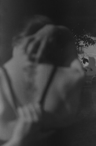 A blurred, monochromatic photograph depicts a figure with indistinct features. They are holding their head with one hand, suggesting a sense of contemplation or distress. In the background, there are out-of-focus foliage and branches, adding a natural element to the composition.