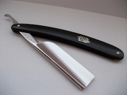 A straight razor with a glossy black handle and shiny metal blade, resting on a smooth, light-colored surface.