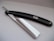 A straight razor with a glossy black handle and shiny metal blade, resting on a smooth, light-colored surface.