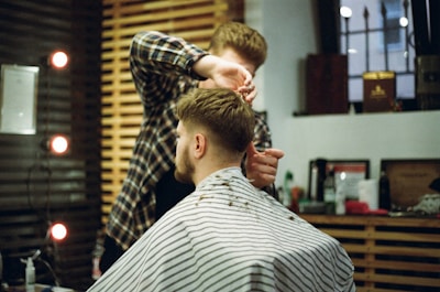 A man in a barber shop chair is having his hair cut by a barber. The barber, wearing a plaid shirt, carefully trims the man's hair. The salon features wooden walls and shelves with various hair products, accompanied by warm lighting from small, circular lights.