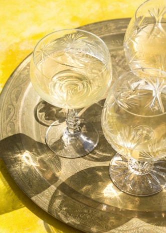 Several crystal glasses filled with a light-colored beverage are placed on an ornate metal tray. The background features a vibrant yellow surface, creating a sunny and cheerful ambiance. The glasses have intricate star-like patterns etched into them, reflecting the light beautifully.
