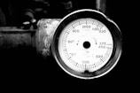 A close-up view of an industrial pressure gauge displaying measurements in a circular format, with the needle indicating a specific pressure reading. The gauge is connected to a weathered metallic surface, showing signs of rust and wear.