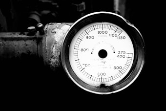 A close-up view of an industrial pressure gauge displaying measurements in a circular format, with the needle indicating a specific pressure reading. The gauge is connected to a weathered metallic surface, showing signs of rust and wear.