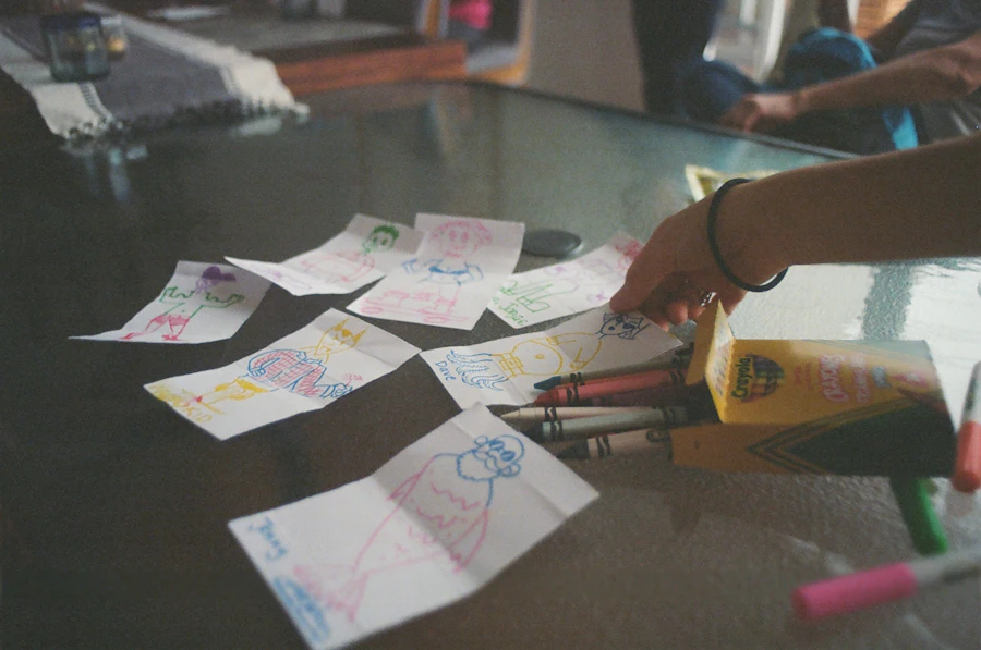 A child's hand reaching across a table covered in crayon drawings on loose sheets of paper