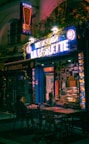 An urban street scene depicting a small kebab shop with bright neon signs that read 'Kebab' and 'La Goulette.' Two people are seated outside at a table, seemingly engaged in conversation while eating. The setting is dimly lit, with a cozy and inviting ambiance created by colorful lighting and foliage surrounding the storefront.