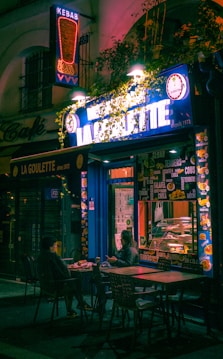 An urban street scene depicting a small kebab shop with bright neon signs that read 'Kebab' and 'La Goulette.' Two people are seated outside at a table, seemingly engaged in conversation while eating. The setting is dimly lit, with a cozy and inviting ambiance created by colorful lighting and foliage surrounding the storefront.