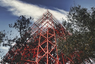 A towering red steel structure with industrial design is surrounded by trees on a partly cloudy day. The intricate lattice of metal beams creates a striking visual contrast against the natural greenery and the blue sky.