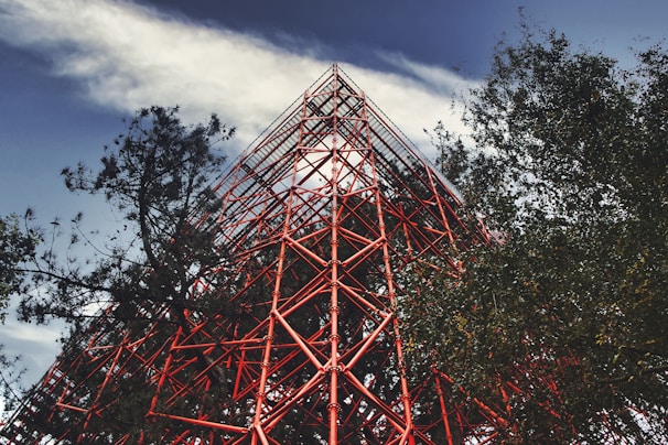 A towering red steel structure with industrial design is surrounded by trees on a partly cloudy day. The intricate lattice of metal beams creates a striking visual contrast against the natural greenery and the blue sky.