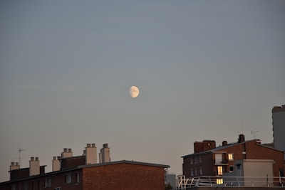 An urban scene featuring several apartment buildings with brick exteriors, some with visible balconies. The sky above them is clear, showing a prominent half moon in the center-right portion of the image.